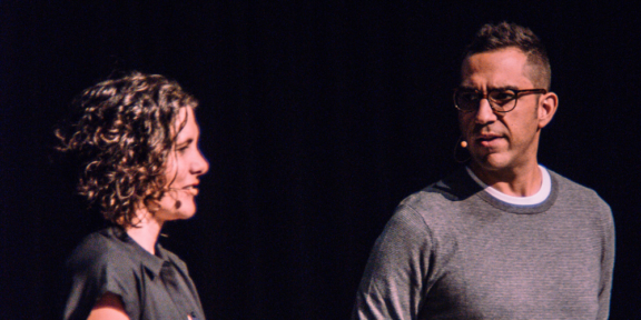 Two presenters wearing headset microphones speak on a dark stage